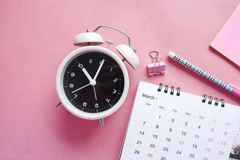 A clock, pen and calendar on a pink table