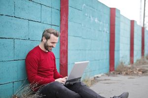 A man with a red long sleeve shirt sitting outside on the ground against a blue wall with his laptop