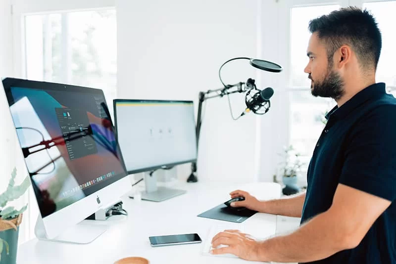 A web designer standing at their desk with two monitors and a podcast microphone