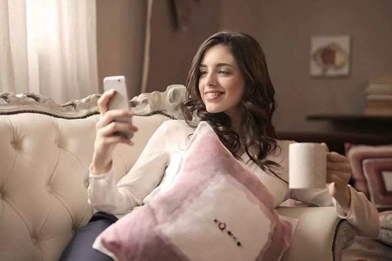 A girl sitting on a sofa with her cup of coffee looking at her mobile phone