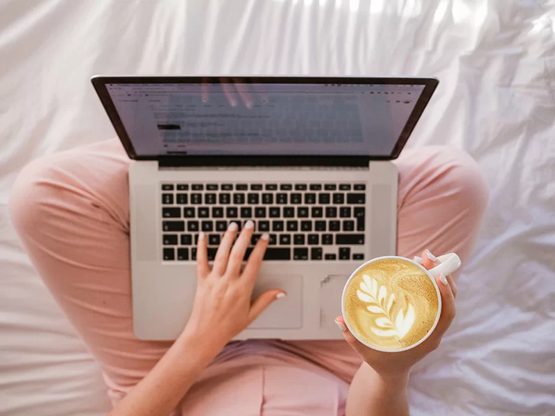 A lady sitting on a bed with a laptop and a coffee in her hand