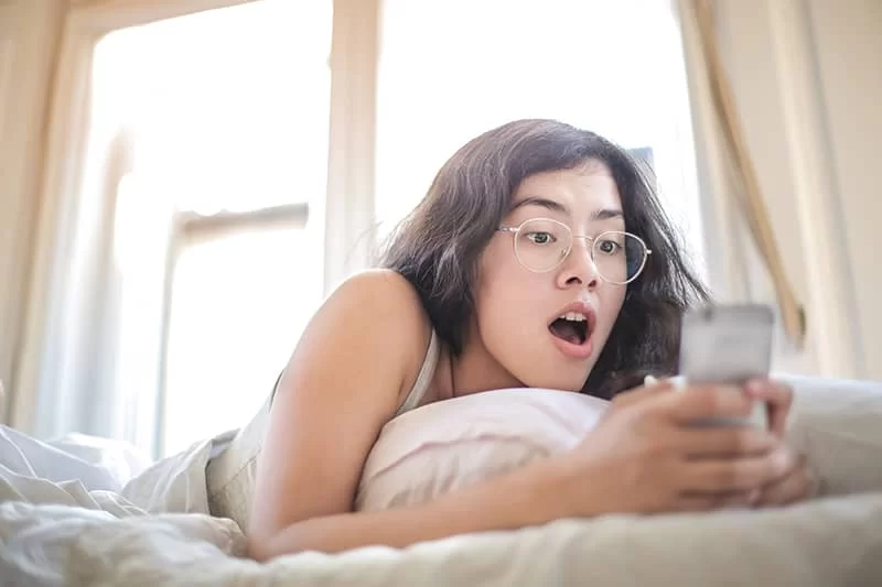 A girl laying on her bed looking at her phone in shock
