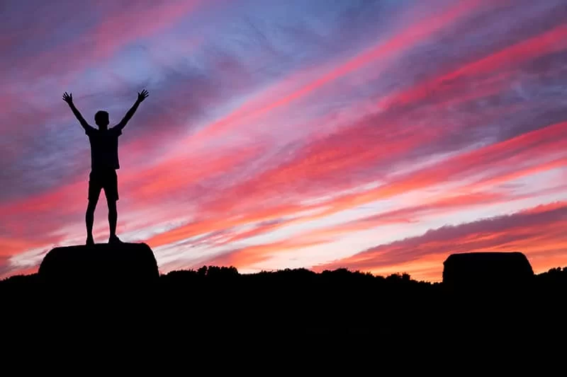 A person celebrating at the top of a hill standing on a rock at sunset