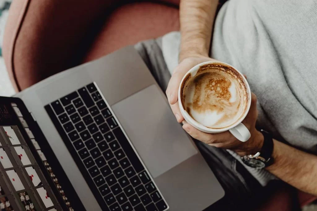 A person sitting in front of a laptop with a cup of coffee in both hands.