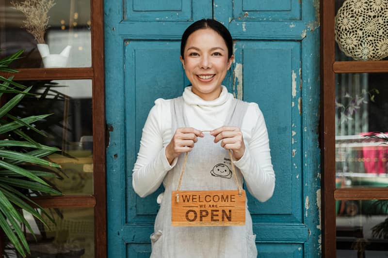 A lady holding a welcome we're open sign in front of her shop